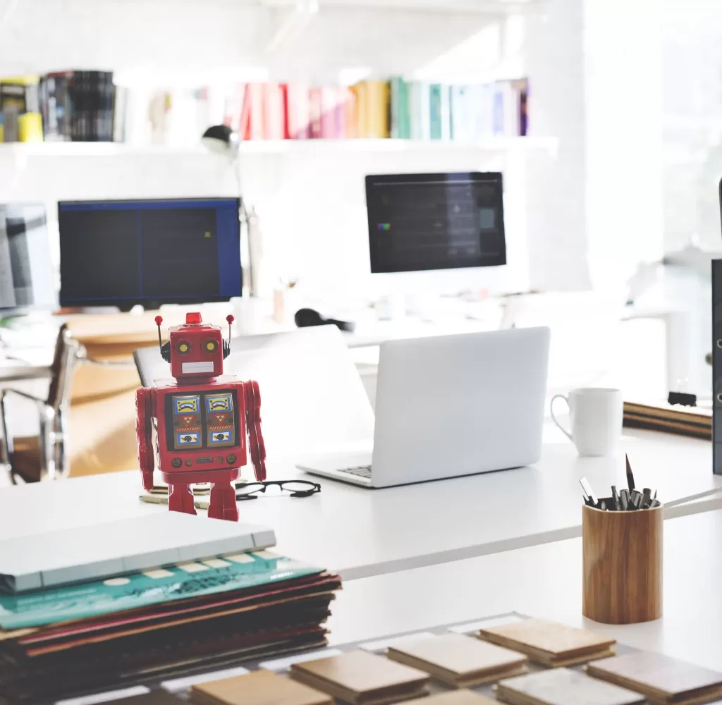 Photo of a desk with a laptop, a robot, and a stack of paper files